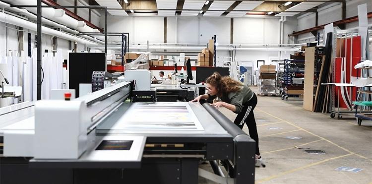 Foto af et printværksted, hvor en kvinde arbejder med en plakat ved en stor printer.